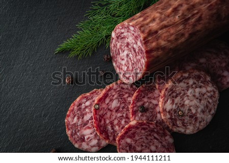 Sliced salami sausage on a board. Meat product and herbs on a dark background. A quick meal or snack. Dried sausage with black peppercorns.