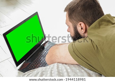 A close-up shot of a Caucasian man lying on the bed and using a green screen laptop