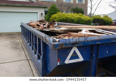 Long blue dumpster full of wood and other debris in the driveway in front of a house in the suburbs Royalty-Free Stock Photo #1944135457