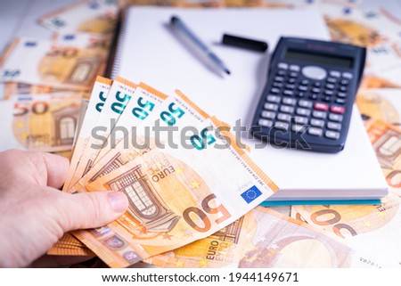 A closeup shot of 50 Euro bills, a calculator and an empty notebook on a wooden surface