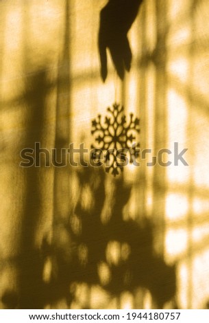 Christmas blurred background with shadow female hand and snowflake christmas toy, abstract aesthetic shadow on yellow textured wall. Sunset and light from window in dark room, at home, real life