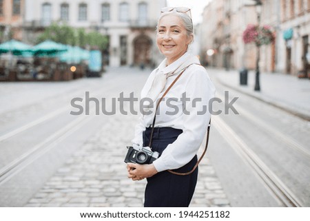 Smiling senior woman in stylish clothes holding retro photo camera while standing outdoors. City street on blurred background. Active lady on retirement.