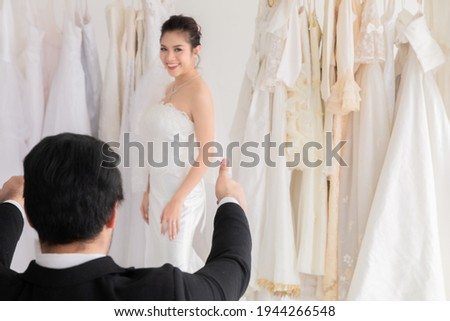 Happy mix race wedding concept, Caucasian groom with beard  sitting to help bride fitting choosing wedding dress in studio, happy beautiful Asian girl show her lace wedding dress to her man
