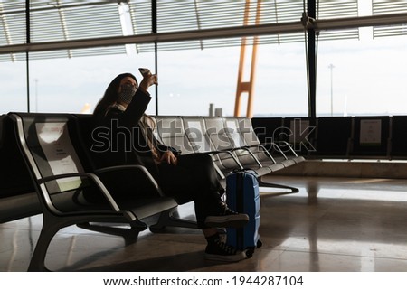 Beautiful young woman taking a picture of himself at the airport. She is sitting on a bench wearing a black and white protective mask. She is carrying a blue suit case. Caucasian and dark haired.