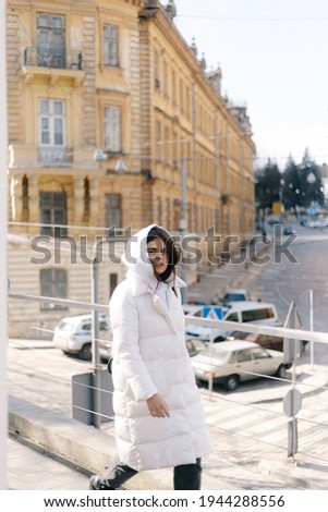A girl in a white jacket and a hood walks along the sidewalk against the background of ancient architecture