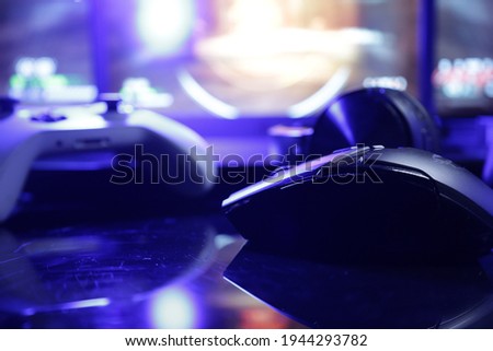 A gaming desk setup with screens illuminating a gamepad and mouse and a gaming laptop on the background with lit keyboard keys