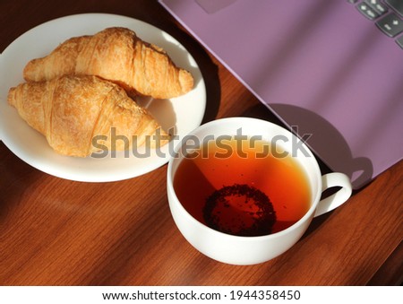 White cup with black tea and croissants in a saucer on the work table. Remote work at home. Snack near the laptop. Sweet morning.