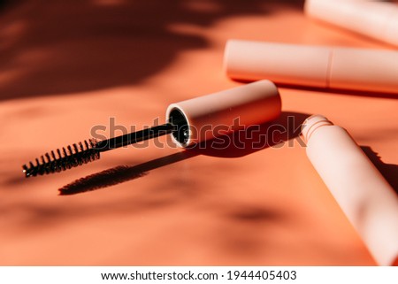 pink clean makeup brush for mascara lies next to an open tube, closed tubes of cosmetics, lip gloss, liquid lipstick, pink eyeliner on a peach background with shadows. Copy space Royalty-Free Stock Photo #1944405403