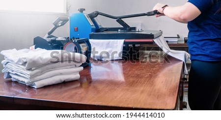 woman manipulating a sublimation machine ironing a shirt. advertising and screen printing concept. Royalty-Free Stock Photo #1944414010