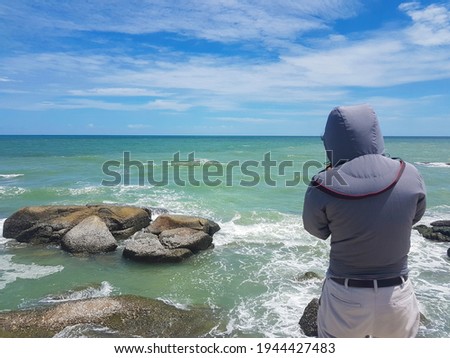 A man in a hooded shirt stands on a rock taking pictures by the sea during the day.