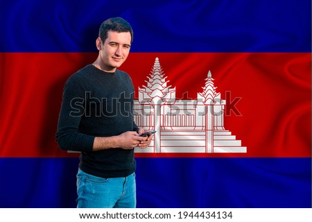 Cambodia flag on the background of the texture. The young man smiles and holds a smartphone in his hand. The concept of design solutions.