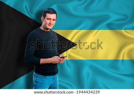 Bahamas flag on the background of the texture. The young man smiles and holds a smartphone in his hand. The concept of design solutions.