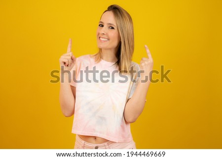 Portrait of a young happy woman dressed in summer clothes pointing fingers up at copy space isolated over yellow background