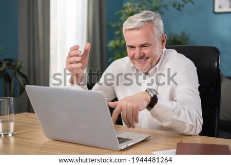 Portrait of a happy smiling guy in his 50s working on a laptop in the living room at home. A handsome gray-haired man looks into the camera and smiles. A fulfilled businessman.