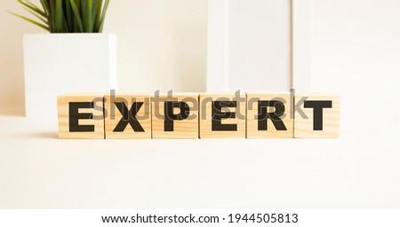 Wooden cubes with letters on a white table. The word is EXPERT. White background with photo frame, house plant.