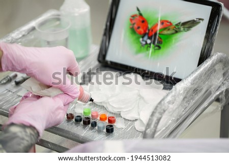 Tattoo master dips a tattoo machine with needles in black ink. The tattoo master applies a colored tattoo on the skin of a young girl, in the background is a tablet with a picture of a ladybug