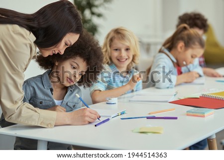 Attentive little schoolboy listening to female teacher while sitting at the table in elementary school classroom together with other kids