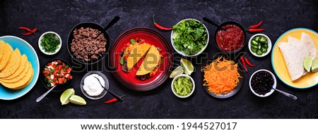 Taco bar table scene with a selection of ingredients. Top view on a dark slate banner background. Mexican food buffet.