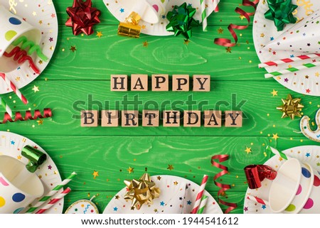 Top view photo of birthday party composition wooden cubes labeled happy birthday ribbon stars spiral candles pipes straws hats sequins paper cups and plates on isolated green wooden table background