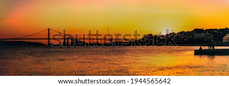 A panoramic shot of a dramatic sunset on the Tagus river with silhouettes of a suspension bridge "25 of Abril" in the background, orange reflections of the sun in the water, houses, and cranes