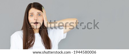 Shocked amazed young woman with hands on head standing and shouting over gray background