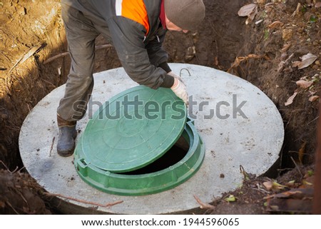 A worker installs a sewer manhole on a septic tank made of concrete rings. Construction of sewerage networks for country houses. Royalty-Free Stock Photo #1944596065