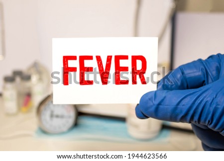 Doctor's hands in blue gloves holding a card with text FEVER on wooden table with stethoscope and notepad for medical records. Medical and healthcare concept. Selective focus