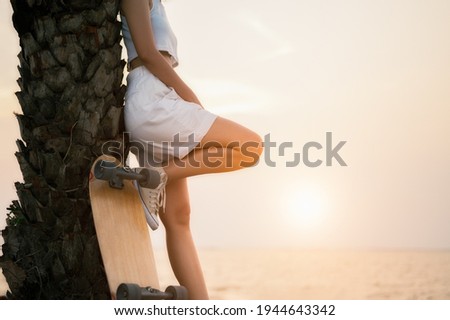 Young women wearing short pants and singlet white leaning against a steel pole with a surf skate next to it. Young woman stand and relax after play surf skate board at skate ramp park on morning.