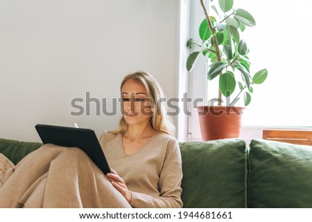 Beautiful blonde young woman artist illustrator drawing on tablet near window at the home