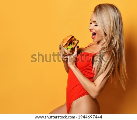 Excited happy screaming slim blonde woman in red swimsuit stands sideways holding big burger in hand and looking inside it, what it was made of. Fast or junk food concept. Girl hold burger