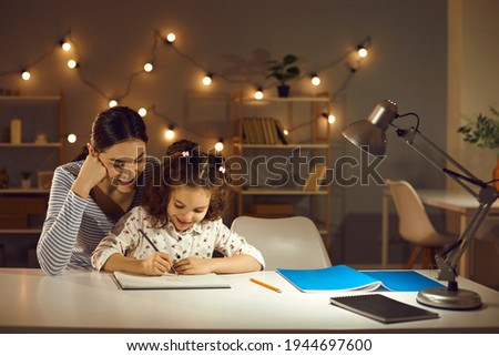 Parent helping child. Happy family doing homework in the evening. Mother and daughter working on school assignment sitting at desk with lamp in cozy dark room with LED lights. Kids learning concept Royalty-Free Stock Photo #1944697600