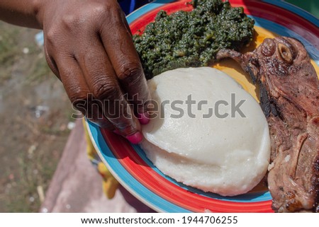 Hand serving on a plate containing African corn meal pap staple food, pumpkin leaves stew and grilled pork Royalty-Free Stock Photo #1944706255