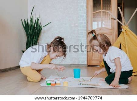 Two children in light clothes sit on the floor on their knees and enthusiastically draw with bright watercolors.