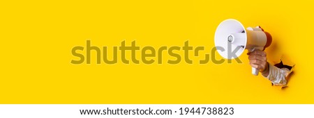 Hand holds a megaphone from a hole in the wall on a yellow background. Concept of hiring, advertising something. Banner. Royalty-Free Stock Photo #1944738823