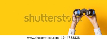 female hands hold black binoculars on a bright yellow background. Banner. Royalty-Free Stock Photo #1944738838