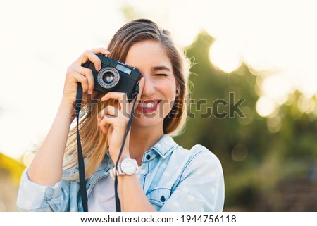 Pretty girl taking photos with a vintage camera on a sunny day, holding it near her eye, smiling. She wears blue sky denim shirt with white big dots and watch. Positive people discover the world.