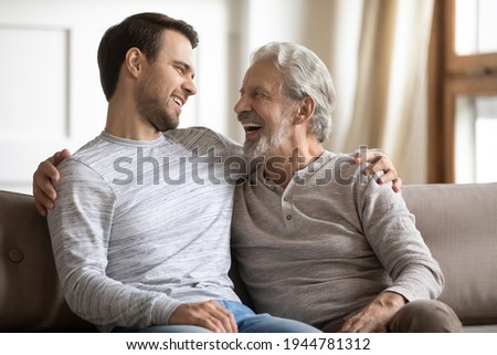 Smiling Caucasian older father and grownup son sit on couch at home hug embrace show love and care in family relations. Happy mature dad and adult millennial man child enjoy reunion on weekend. Royalty-Free Stock Photo #1944781312