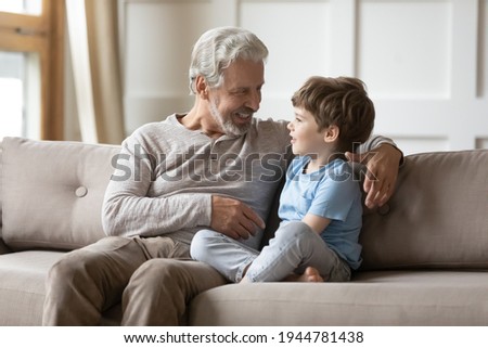 Loving old 60s Caucasian granddad sit on sofa with little curious grandson talk and chat at home. Caring mature grey-haired grandfather relax on couch with small grandchild gossip share secrets. Royalty-Free Stock Photo #1944781438