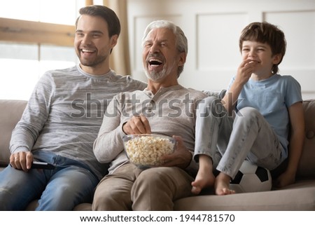 Overjoyed three generations of Caucasian men sit relax on sofa at home laugh watching video eating popcorn together. Smiling boy child with young dad and grandfather rest with snacks enjoy TV.