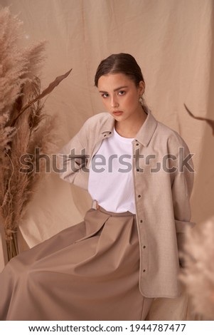 Young elegant female model sitting at beige textile background and posing between ears of rye. Brunette girl with makeup and pony tail hair in white shirt and beige skirt and jacket