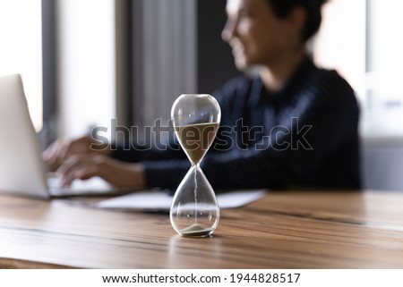 Close up hourglass measuring time, standing on wooden office table, Indian businesswoman working on background, efficiency, deadline and time management concept, busy employee using laptop Royalty-Free Stock Photo #1944828517