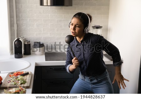 Close up funny Indian woman wearing headphones singing into kitchenware as microphone, dancing to favorite music in modern kitchen at home, overjoyed young female having fun, preparing meal Royalty-Free Stock Photo #1944828637