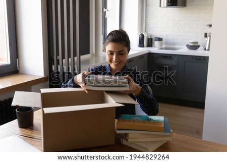 Smiling Indian woman unpacking parcel, holding books, sitting at table, satisfied customer received online store order, delivery service, happy female packing belongings, preparing for relocation Royalty-Free Stock Photo #1944829264