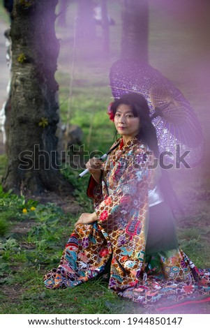 A beautiful Japanese girl in a kimono with floral pattern sits in a blossoming garden and looks through flowers on camera. A woman in traditional Japanese clothes sits with a violet umbrella.