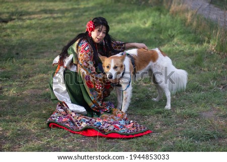 A beautiful woman in a flower kimono sits together with a dog and looks on camera. A model in traditional colorful Japanese clothes and dog on green background. Warm connection between close friends. 