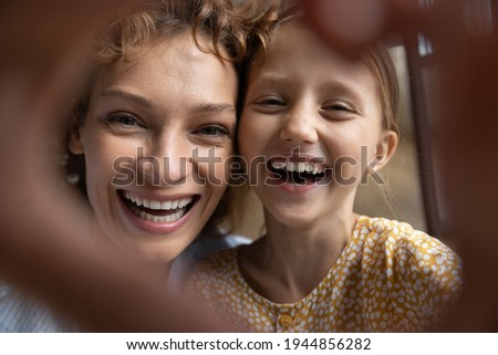 Close up of overjoyed young Caucasian mother and little 9s daughter have fun make selfie together. Smiling mom and small girl child laugh take self-portrait picture show heart love hand gesture.