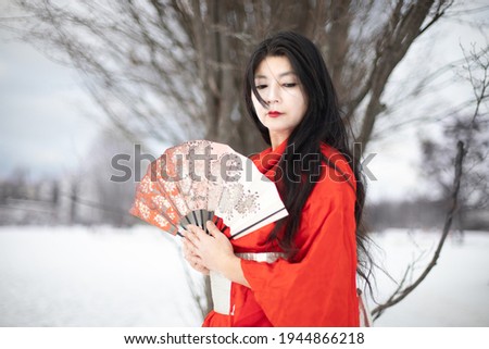 A breathtaking picture. A professional portrait of a pretty Japanese girl with a white face who stands outdoor on a cold day and looks down. A woman in traditional red clothes with a fan in her hands.