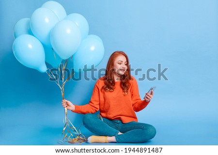 European woman sitting on floor, holding bunch of helium balloons, female with phone looking at display of device, calling somebody to invite to her party.