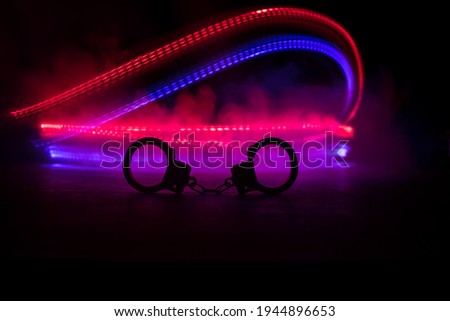 Police raid at night and you are under arrest concept. Silhouette of handcuffs with police car on backside. Image with the flashing red and blue police lights at foggy background. Selective focus