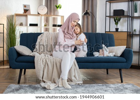 Happy motherhood, Mother day concept. Pretty young smiling Muslim woman mother hugging cute little daughter, posing to camera at home in cozy living room. Mom and child in domestic wear